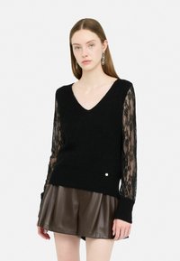 Maglione nero a V con corpo a maglia strutturata e maniche lunghe in pizzo trasparente. Presenta un piccolo dettaglio dorato all'orlo. Abbinato a pantaloni corti in pelle marrone.