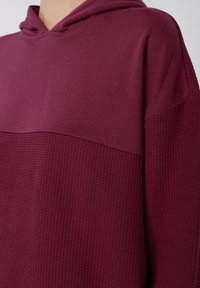 Sweatshirt à capuche cramoisi présentant une partie supérieure lisse et un panneau inférieur texturé avec un motif en grille subtil. Convient pour une tenue décontractée.