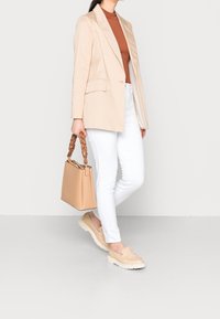 Beige op maat gemaakte blazer over een bruine top, witte slim-fit broeken en beige loafers, met een tan leren tas met een gevlochten handvat.