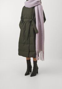 Olivově zelený prošívaný kabát s páskem v pase, doplněný světlým levandulovým šátkem a černými botami na podpatku, s texturovaným povrchem.