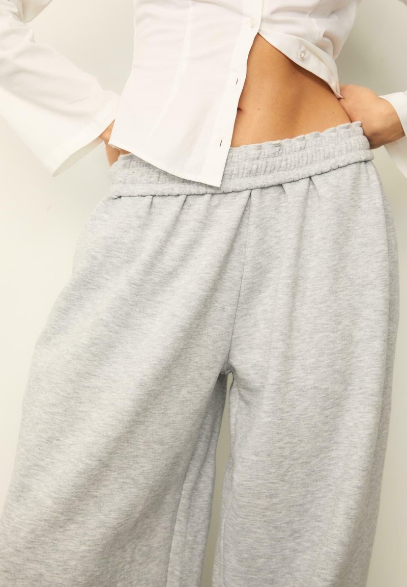 Graue Baumwoll-Sweatpants mit elastischem Bund, lockerer Passform und subtiler Textur. Kombiniert mit einem weißen Hemd mit Knöpfen.