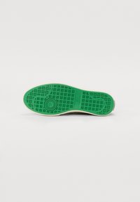 Semelle de chaussure en caoutchouc vert avec un motif de grille texturé, entourée d'une bordure crème, dotée d'un détail de logo circulaire au niveau du talon.