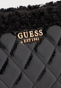 Čierna prešívaná kabelka s nadýchaným čiernym textilným lemom. Zlaté logo "GUESS EST 1981" je výrazne zobrazené na prednej strane.