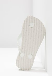 Hvid flip-flop sandal, der læner sig mod en hvid hjørnevæg og viser tekstureret sål med tre cirkulære propper.