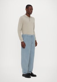 Beige Langarm-Polohemd kombiniert mit hellblauen weiten Hosen und schwarzen Schuhen. Das Polohemd hat einen Kragen und ist an den Handgelenken eng geschnitten.