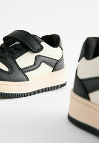 Sorte og creme lav-top sneakers med en tekstureret læderoverflade. Har en sikker rem og gummisål med et mønstret dæk.
