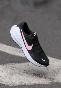 Μαύρο αθλητικό παπούτσι Nike με ροζ λογότυπο swoosh και λευκή σόλα, αιωρούμενο στον αέρα πάνω από επιφάνεια από άσφαλτο.