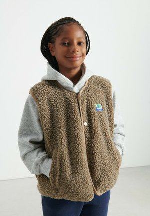 Braune Fleeceweste mit strukturierter Oberfläche und beigem Besatz, über einem grauen Kapuzenpullover getragen, mit buntem Markenpatch und Frontknöpfen.