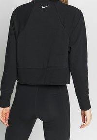 Persona vestida con sudadera negra de manga larga cortada con el logo blanco de Nike en la parte superior de la espalda y leggings negros ajustados, vista desde atrás.