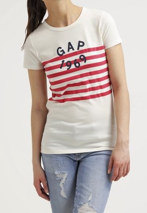 Valkoinen lyhythihainen t-paita, jossa on punaisia vaakaraitoja ja laivastonsininen teksti "GAP 1969" rinnassa. Yhdistetty sinisiin kuluneisiin farkkuihin.