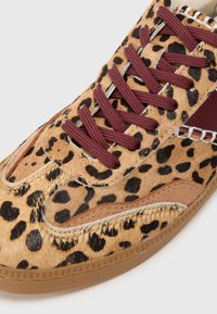 Tenis cu imprimeu leopard, cu partea superioară texturată de culoare bej, sireturi vișinii și talpă din cauciuc. Coaserea notabilă și accentele de panou în culori contrastante.