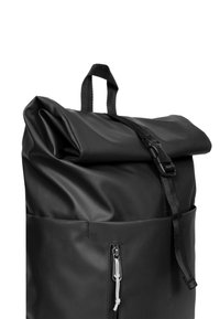 Sac à dos noir à rabat fabriqué dans un matériau durable, doté d'une poignée supérieure, d'une sangle réglable et d'une poche avant zippée avec tirette blanche.