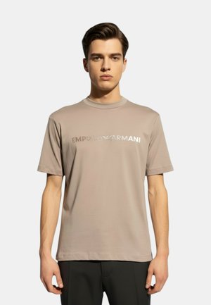 Giovane uomo con capelli scuri che indossa una t-shirt beige Emporio Armani con logo metallico, in piedi davanti a uno sfondo chiaro semplice.