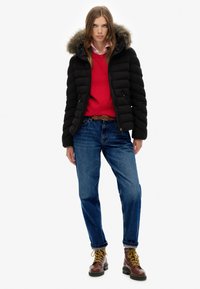 Chaqueta acolchada negra con capucha forrada de piel, suéter rojo debajo, vaqueros azules y botas marrones con cordones amarillos; conjunto informal.