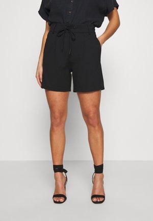 Femme portant un short noir taille haute avec cordon de serrage et poches, associé à une chemise noire boutonnée et des talons noirs attachés à la cheville, debout.