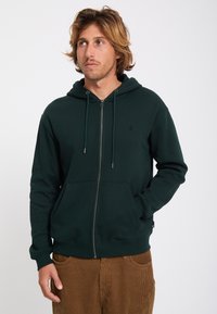 Jeune homme aux cheveux ondulés portant un sweat à capuche zippé vert foncé et un pantalon en velours côtelé marron, regardant légèrement sur le côté.
