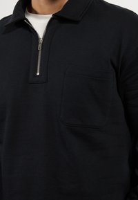 Hombre vestido con una camiseta de manga larga negra con cremallera de un cuarto y un bolsillo en el pecho, sobre una camiseta blanca interior, mostrado desde el cuello hasta la mitad del torso.