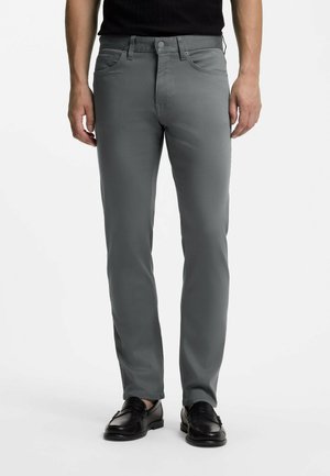 Mann trägt eng anliegende graue Jeans kombiniert mit schwarzen Lederloafers und einem schwarzen Hemd, steht vor einem einfarbigen weißen Hintergrund.