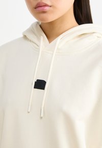 Cremefarbene Hoodie mit Kordelzug, ausgestattet mit einem schwarzen Logopatch. Der Stoff wirkt weich mit einer glatten Textur und hat eine reguläre Passform.