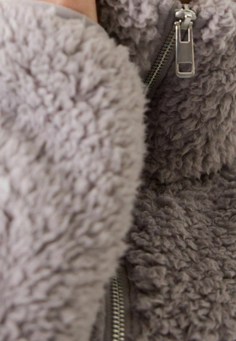 Gros plan d'une veste en tissu gris, duveteux et texturé, avec une fermeture éclair argentée visible, montrant un matériau doux et épais.