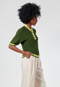 Green knitted polo shirt with a yellow collar and trim, featuring a textured herringbone pattern and four buttons down the front.