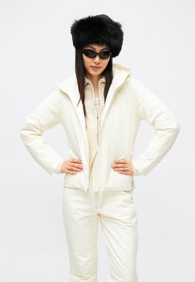 Fehér, kapucnis bélelt dzseki krémszínű pulóverrel és fehér nadrággal párosítva. Kiegészítve fekete szőrmekalappal és fekete napszemüveggel.