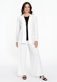 Blazer blanc à manches longues, fermeture à boutons, associé à un pantalon large blanc, un top noir en dessous et des sandales à talons noires.