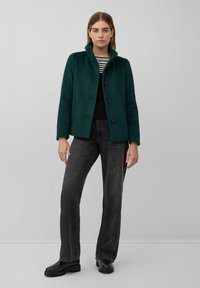 Grüner Wollmantel mit Stehkragen, Knopfverschluss, einer Brusttasche, getragen über einem schwarzen Strickoberteil und dunkelgrauen Jeans, kombiniert mit schwarzen Stiefeln.