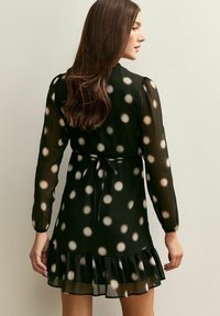 Zwarte jurk met stippen, lange mouwen, een rucheszoom en een tailleband, gemaakt van doorschijnende stof met een zachte textuur.