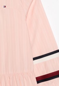 Vaaleanpunainen mekko, jossa on tekstuuripinta, pitkät hihat ja nauhakuviot tummansinisessä, valkoisessa ja burgundissa hihojen resoreissa.