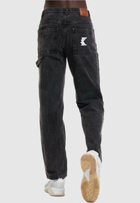 Black denim jeans with a relaxed fit, featuring back pockets and a white embroidered logo on the right pocket. Pairing with white sneakers.