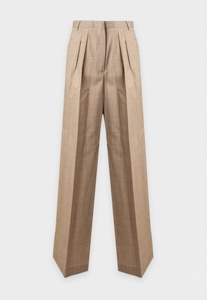 Femme portant une veste en cuir marron sur un haut blanc boutonné et un pantalon ample beige à jambes larges, debout les mains dans les poches.