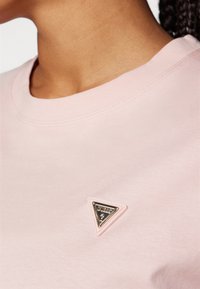 T-shirt de algodão rosa claro com decote redondo, apresentando um logotipo metálico triangular em uma moldura elevada no peito.
