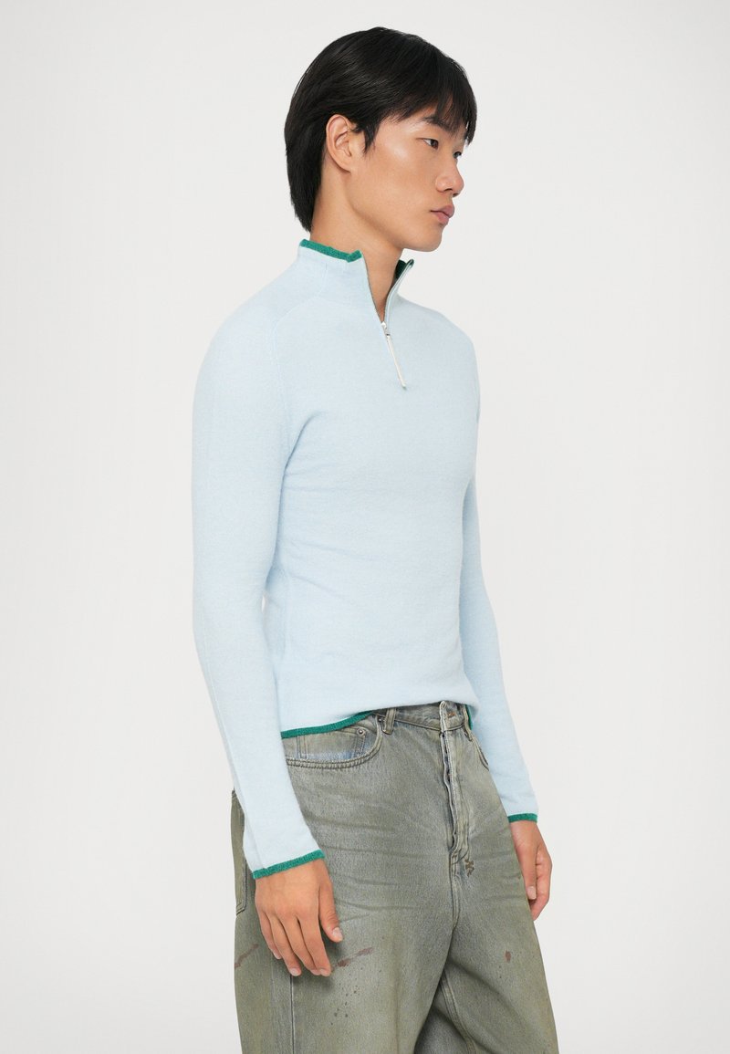 Ljusblå zip-up tröja med gröna detaljer vid kragen och ärmsluten, figursydd design, mjuk textur, i par med ljusgrå baggy jeans.