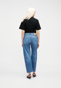 Ξανθιά γυναίκα που φοράει μαύρο T-shirt, μπλε ψηλόμεσο τζιν και μαύρα παπούτσια με τακούνι, στεκόμενη πλάτη σε απλό λευκό φόντο.