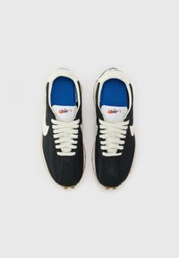 Crne mrežaste tenisice s bijelim vezicama, gumiranim potplatom i plavim ulošcima. Nike logo na jeziku i peti, s teksturiranim dizajnom.