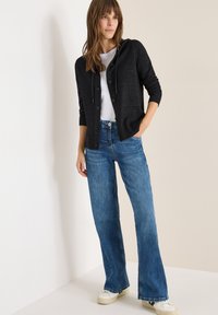 Schwarze Strickjacke mit Kapuze, darunter ein weißes Hemd, kombiniert mit blauen Schlagjeans aus Denim und weißen Turnschuhen vor einem neutralen Hintergrund.