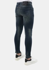 Slim-fit donkerblauwe denim jeans met vervagingdetails, vijf zakken en een zwartleren label op de taille, gecombineerd met witte sneakers.