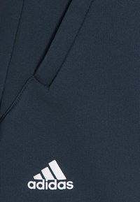 Tmavě modré sportovní kalhoty vyrobené z texturované látky, s bílým logem Adidas v dolním levém rohu a boční kapsou na úložný prostor.