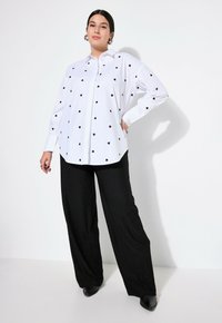 Witte button-up blouse met zwarte hartpatronen, gecombineerd met zwarte wijde broek. De blouse heeft een klassieke kraag en lange mouwen.