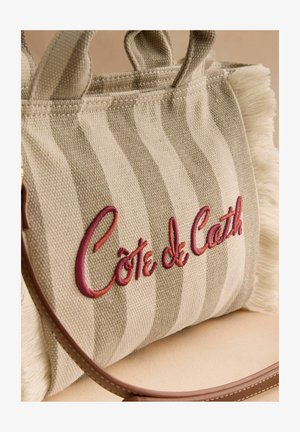 Beżowo-kremowa paskowana torba materiałowa z frędzlami na brzegach i czerwonym haftem "Côte de Cath" po boku.