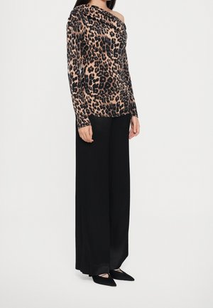 Femme portant un haut léopard à manches longues et épaules dénudées, avec un pantalon noir à jambes larges et des chaussures noires pointues, debout sur fond blanc.
