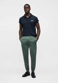 Άνδρας όρθιος με τα χέρια στις τσέπες, φοράει μαύρη πόλο μπλούζα, πράσινο αθλητικό παντελόνι και μαύρα αθλητικά παπούτσια μπροστά σε απλό λευκό φόντο.