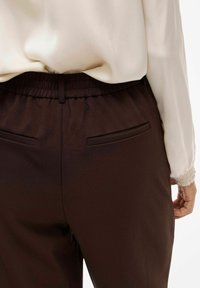 Pantaloni con fascia elastica marrone e tasche, realizzati in tessuto liscio. La parte superiore è di colore crema, con un polsino delle maniche testurizzato visibile.