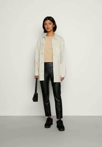 Camisa de botão oversized às riscas em branco e azul marinho, top bege, calças de pele sintética pretas, sapatilhas pretas e uma pequena bolsa preta.