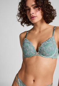 Groene bloemenkant beha met gekartelde randen, verstelbare schouderbanden en bloemenborduursel in paarse en witte accenten, versierd met een klein strikje.