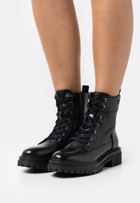 Botas de combate de cuero negro con punta redonda, cierre de cordones en la parte delantera, cremallera lateral, suela de goma gruesa y acentos de hardware plateado.