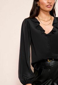Black blouse with ruffled neckline and long, puffed sleeves. Smooth fabric and button front. Worn with black leather pants and layered gold necklaces.