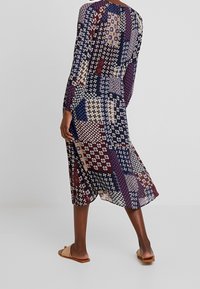 Femme portant une robe midi à manches longues au motif patchwork en marine, bordeaux, beige et blanc, associée à des sandales plates beiges à bout ouvert.