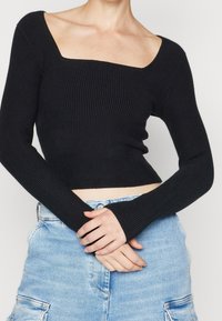 Sort ribstrikket langærmet top med firkantet udskæring, tætsiddende design og tommelfingerhul-detaljer. Båret over lyse denimshorts.